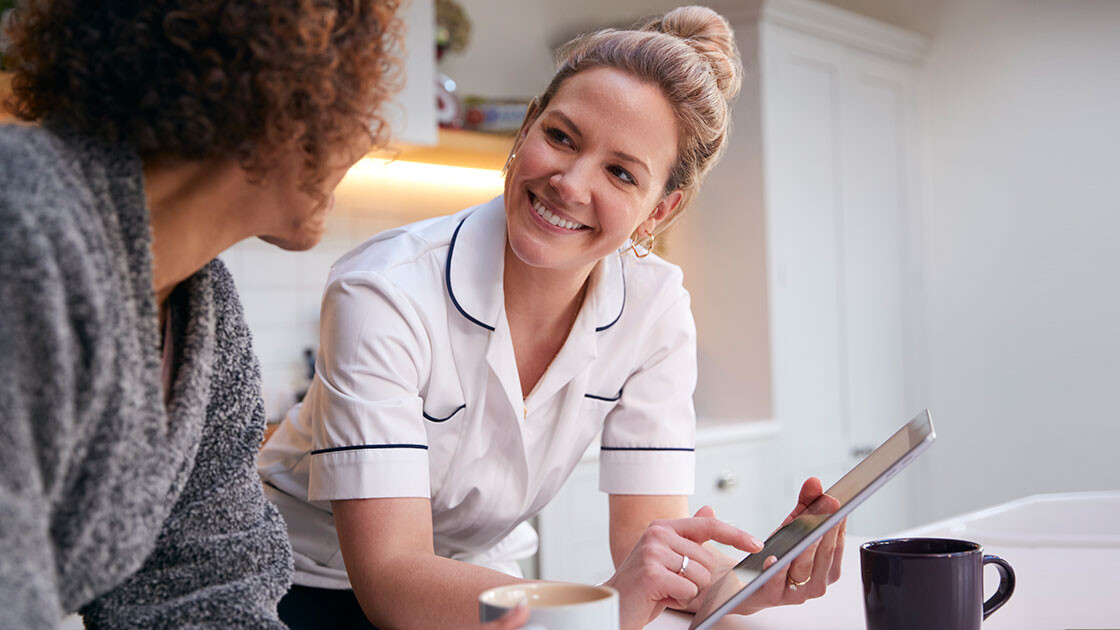 Nurse smiling while using a tablet to review care information with a long-term care resident in a warm, comfortable setting.
