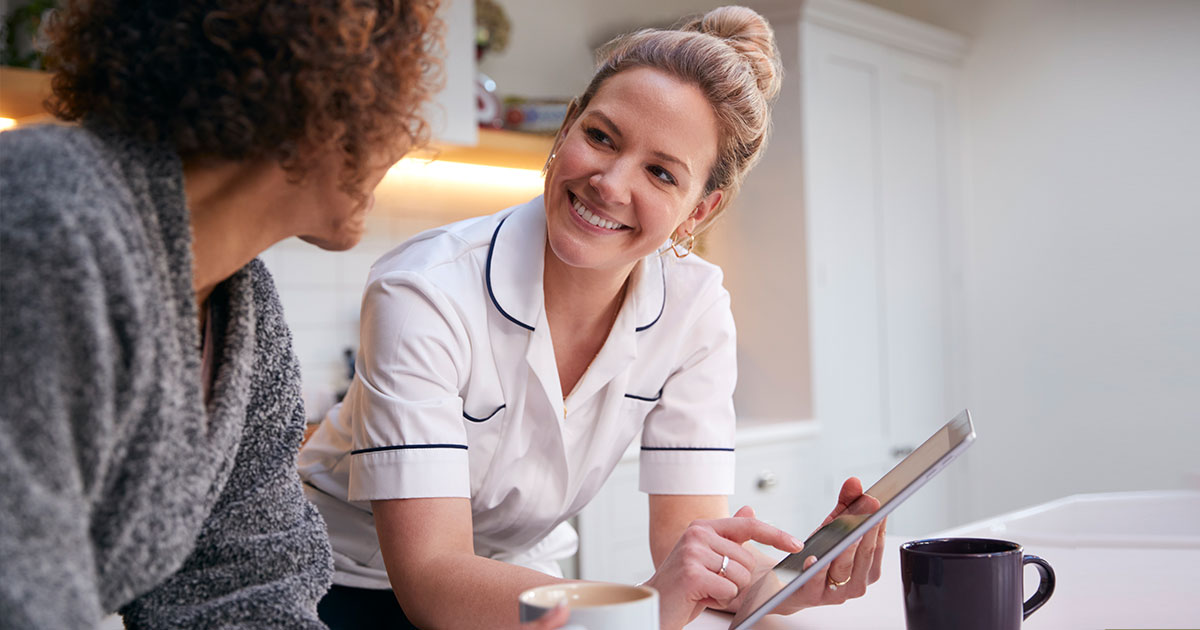 Nurse smiling while using a tablet to review care information with a long-term care resident in a warm, comfortable setting.